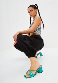 Sandales à talons blocs vert menthe avec une finition brillante, présentant un bout carré et un design à brides croisées. Associées à un pantalon noir texturé.