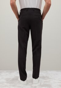 Zwarte broek met een slim fit, voorzien van een platte tailleband, twee achterzakken met knopen en een gladde textuur. In combinatie met witte sneakers.