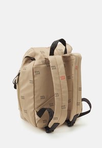 Zaino in tessuto beige con motivo logo nero, doppie spalline nere e chiusura con zip. Gli accenti includono un dettaglio logo rosso a contrasto.
