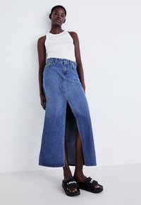 Jupe maxi en denim avec une fente avant, présentant une couleur bleu délavé et des coutures contrastées. Associée à un débardeur blanc et des sandales noires.