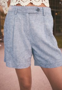 Světle modré lněné šortky s texturovaným povrchem, se sklady, předním zapínáním na knoflík a bočními kapsami. Lemování je mírně ohrnuté.