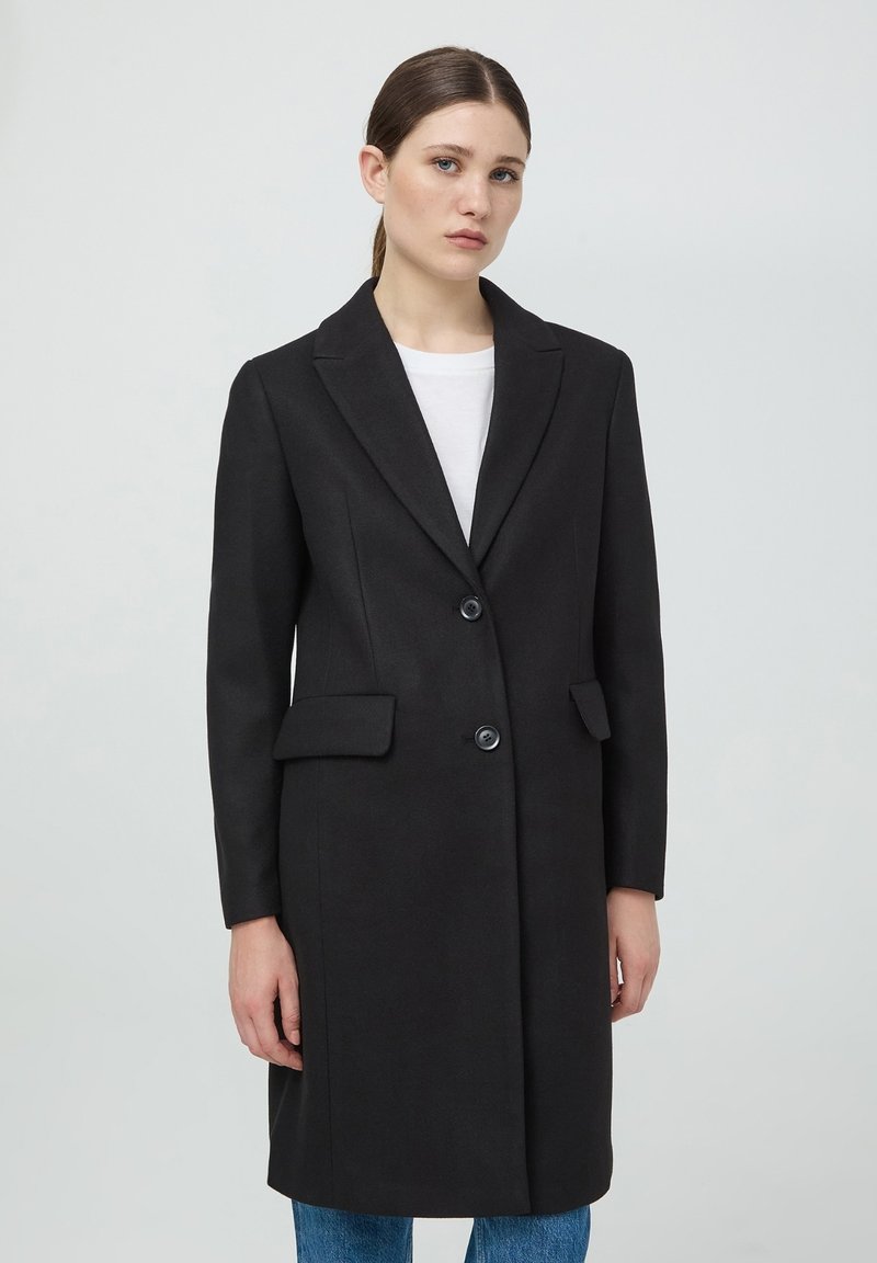 Cappotto in misto lana nero con revers, due bottoni, tasche laterali e silhouette sagomata. Indossato su una maglietta bianca e jeans blu.