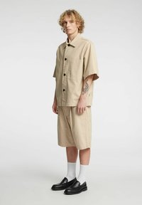 Beige skjorte med knapper, korte ermer og en lomme, kombinert med matchende knelang shorts. Svarte lær loafers og hvite ankelsokker.