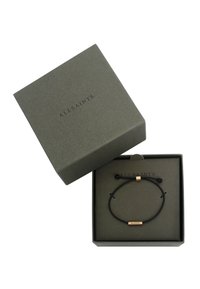 Bracciale di corda nera con un accento metallico dorato su cui è inciso "ALLSAINTS", presentato in una scatola quadrata grigia texturizzata.