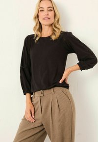 Blonde Frau, die ein schwarzes Langarmoberteil mit Spitzenakzenten und hochgeschnittene beigefarbene Faltenhose trägt, steht mit der Hand auf der Hüfte.