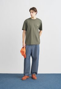 Λαδί βαμβακερό t-shirt με κοντά μανίκια, συνδυασμένο με άνετο μπλε τζιν παντελόνι. Αξεσουάρ περιλαμβάνουν πορτοκαλί καπέλο και αντίστοιχα παπούτσια.