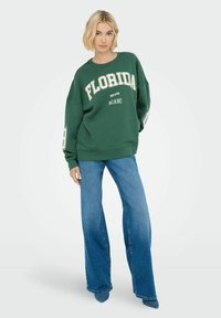 Felpa verde con la scritta "Florida" e "Miami" in crema. Abbinata a jeans a gamba larga blu e tacchi a punta blu.