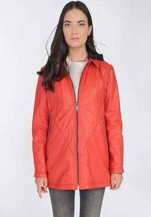 Vrouw draagt een rode leren jas met rits over een licht shirt en donkere spijkerbroek, staand tegen een effen lichte achtergrond.