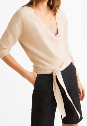 Vrouw draagt een beige wikkeltrui met ceintuur en zwarte knielange shorts met verticale strepen, poseert met één hand in de zak.