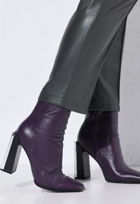 Fialové kožené kotníkové boty s volným designem, špičatou špičkou a robustním černým podpatkem. Stylizováno s šedými koženými kalhotami.