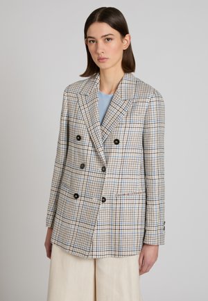 Vrouw draagt een geruit dubbelbreasted blazer met zwarte knopen over een lichtblauw topje en beige wijde broekspijpen.