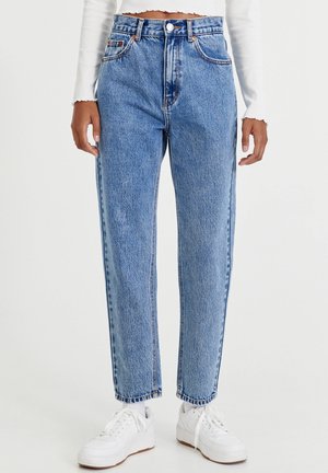 Persoon draagt een blauwe high-waisted jeans met knoop en ritssluiting, gecombineerd met witte sneakers en een witte cropped top met lange mouwen.