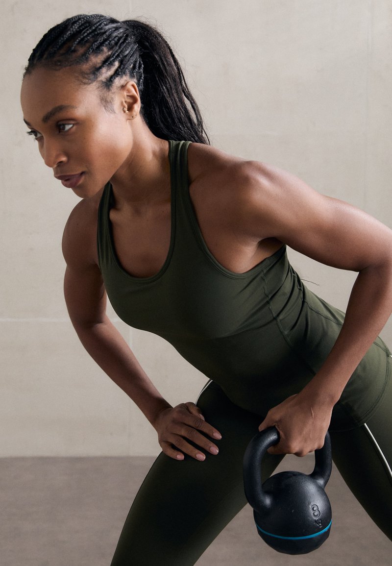 Um kettlebell preto com um detalhe azul é segurado por uma pessoa que veste uma t-shirt de treino justa em verde-escuro e leggings a condizer, mostrando definição muscular.