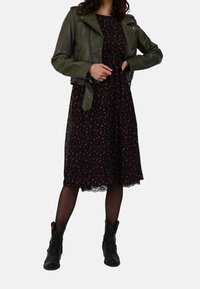 Green leather biker jacket over a black floral midi dress with lace trim, paired with black ankle boots. Simple jewelry accents.