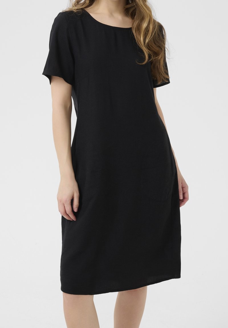 Femme portant une robe noire courte aux manches courtes et au col rond, debout devant un fond clair uni.