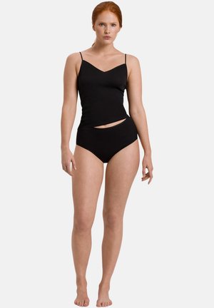 Donna con capelli rossi che indossa una canottiera nera e mutandine a vita alta nere, in piedi davanti a uno sfondo bianco uniforme.