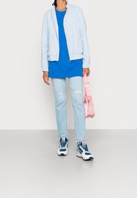 Ljusblå bomberjacka, blå t-shirt, rivna ljusblå jeans och vita sneakers med blå accenter. Håller en rosa genomskinlig väska.