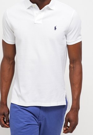Polo shirt bianco in cotone, con una pattina a tre bottoni e un logo blu navy sul lato sinistro del petto. Maniche corte e texture morbida.