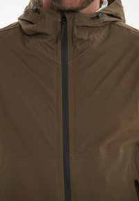 Veste imperméable marron avec capuche, dotée d'une fermeture éclair sur le devant et d'accents noirs le long de la fermeture éclair et des poches latérales. Texture lisse.
