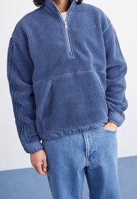 Blå fleece-pullover med halvdragkedja, texturerad yta, elastisk midja och sidofickor. Har kontrasterande tygdetaljer.
