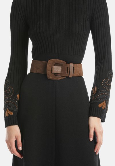 Brauner Wildledergürtel mit einer rechteckigen Schnalle, getragen über einem schwarzen, gerippten Strickpullover mit gestickten Blumendesigns an den Ärmeln.