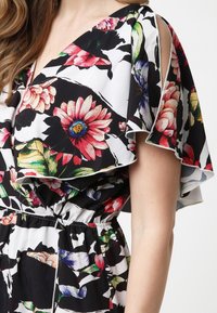 Femme portant une robe fleurie avec des manches volantées et une taille cintrée, avec de grands motifs floraux roses, rouges, violets et verts.