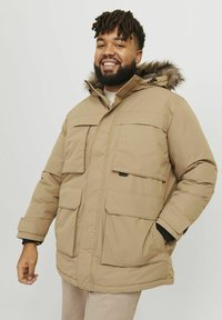 La chaqueta puffer color canela cuenta con una capucha forrada de piel sintética, múltiples bolsillos frontales y un cierre frontal con cremallera. Tejido texturizado con un acabado suave.