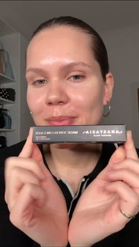 Personne à la peau claire tient une boîte noire portant l'étiquette "Anastasia Beverly Hills Magic Touch Concealer" près de son visage.