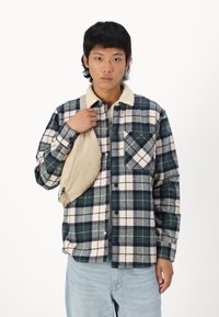Chemise en flanelle à carreaux, verte et blanche, avec un col en sherpa clair et une poche poitrine. Portée avec un sac à bandoulière beige.