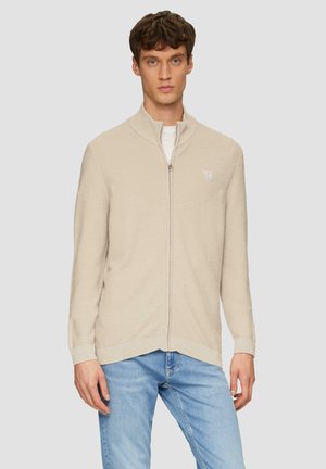 Pull-over zip beige avec un col haut, tissu texturé, design ajusté, et logo discret sur la poitrine gauche, associé à un jean bleu clair.