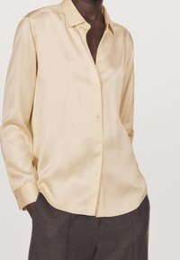 Chemise beige en satin à boutons, avec col, manches longues et texture lisse, partiellement rentrée dans un pantalon sombre et sur mesure.