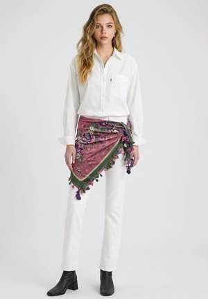 Mujer con camisa blanca abotonada, pantalones blancos, botas negras hasta el tobillo y una bufanda morada con flores atada alrededor de su cintura con flecos.