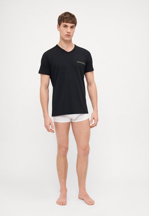 T-shirt nera a scollo a V realizzata in tessuto morbido, con un piccolo logo sul petto. Indossata con slip bianchi, in piedi su uno sfondo neutro.
