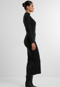 Abito lungo nero a maniche lunghe con un design aderente, realizzato in tessuto morbido. Presenta un collo alto e una fessura laterale, abbinato a stivali neri sopra la caviglia.