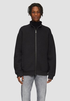 Zwarte zip-up sweatshirt met een gestructureerde afwerking, hoge kraag en geribbelde manchetten. Gepaard met lichtgrijze jeans.
