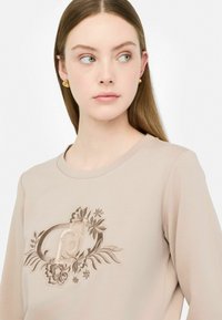 Felpa beige a maniche lunghe con un design di ricamo floreale e un emblema sul petto. Texture liscia e collo a giro.