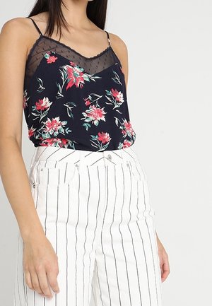 Kobieta ubrana w granatową kwiecistą koszulkę na ramiączkach z koronkowym wykończeniem, w połączeniu z wysokimi białymi spodniami z cienkimi pionowymi czarnymi paskami.