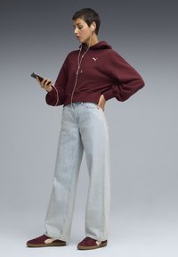Bordeaux cropped hoodie, lyseblå vide jeans og burgundy sneakers. Modellen holder en smartphone og har hvide earbuds på.