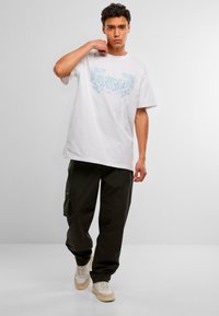 Joven con cabello rizado, vistiendo una camiseta gráfica blanca, pantalones cargo negros y zapatillas beige, de pie frente a un fondo blanco liso.