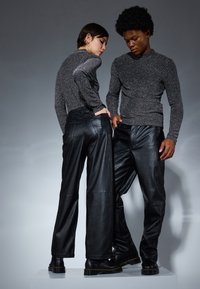 Dos personas que llevan tops de manga larga con brillo y pantalones de cuero negro texturizados, de pie sobre una plataforma blanca contra un fondo gris.