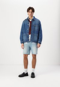Veste en denim bleu moyen, avec fermeture à boutons et deux poches avant. Chemise blanche sous une cravate bordeaux, associée à un short bleu clair et des chaussures noires.