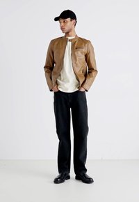 Giacca di pelle marrone con design senza colletto, tasca con zip, abbinata a una camicia chiara, pantaloni neri e scarpe nere su uno sfondo neutro.