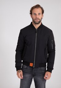 Zwarte bomberjack met ritssluiting, geribde manchetten, zijzakken en een opvallend oranje label aan de onderkant. Gladde stoftextuur.