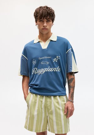 Polo atletico blu e crema a maniche corte, con testo e loghi, abbinato a pantaloni corti a righe verdi chiare e bianche.