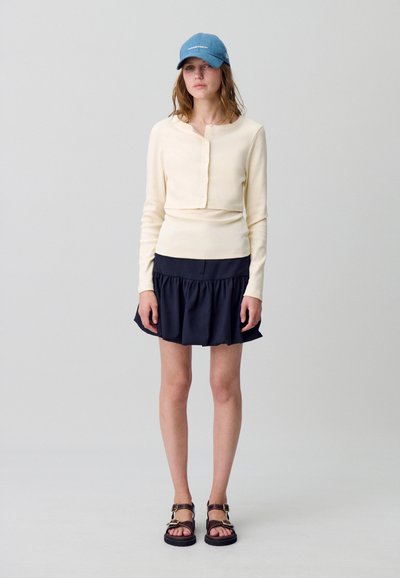 Femme portant une casquette bleue, un haut à boutons crème à manches longues sur une mini-jupe noires à volants, et des sandales marron, se tenant devant un fond uni.