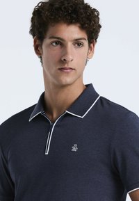 Polo shirt bleu marine avec un col zippé, des accents blancs et un petit logo brodé sur la poitrine. Le matériau est un mélange de coton texturé.