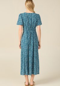 Blumenkleid in Taubenblau aus einem leichten Stoff mit kurzen Ärmeln, elastischem Bund und weißem Blumenmuster. Der Rücken ist mit einem gerafften Detail versehen und der Rock ist fließend.