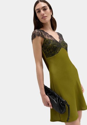 Kvinde iført olivengrøn kjole med sort blonde topdetalje, der holder en sort ruched clutch taske mod en ensfarvet baggrund.