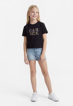 Fată zâmbitoare stă în picioare purtând un tricou negru cu logo Emporio Armani, pantaloni scurți ușori din denim și adidași albi pe fundal alb.