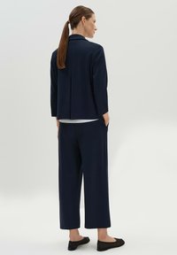 Navyblauer maßgeschneiderter Blazer und weit geschnittene Hose aus weichem Stoff. Der Blazer hat einen Reverskragen und einen Rückenschlitz. Schwarze flache Schuhe.